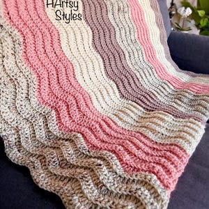 May include: A handmade crochet blanket in a wavy pattern, featuring stripes of pink, cream, and taupe. The blanket is draped over a dark blue surface. The text "HArtsy Styles" is visible on the blanket.