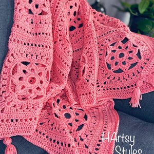 May include: A coral-coloured, handmade crochet blanket with a floral design. The blanket features intricate openwork patterns and is adorned with tassels at the corners. The blanket is draped over a surface, showcasing its texture and detail. The text "HArtsy Styles" is visible in the image.
