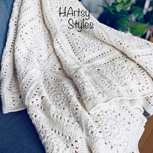Pode incluir: Cobertor de crochê feito à mão, creme, com um padrão floral e geométrico. O cobertor está drapeado sobre uma superfície azul. O texto "HArtsy Styles" é visível.