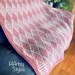 May include: A crocheted blanket featuring a repeating geometric diamond pattern in dusty rose and white yarn. The textile displays a textured stitch design throughout the fabric. The text HArtsy Styles is printed in white in the bottom left corner.