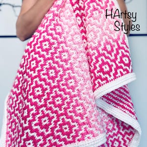 Könnte beinhalten: Eine Häkeldecke mit einem geometrischen Rautenmuster in Rosa- und Weißtönen. Das Design verläuft von tiefem Magenta zu helleren Rosatönen und ist mit einem einfarbigen weißen Rand versehen. Der Text HArtsy Styles ist auf dem Stoff zu sehen.