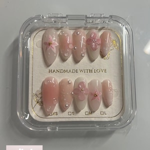May include: A set of 10 pink and white press-on nails with 3D flower accents. The nails are arranged in a clear plastic case with size labels: XS, S, M, L. The case has the text "HANDMADE WITH LOVE" printed on it.