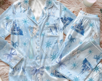 May include: Light blue pyjama set with a princess design, snowflakes, and castle illustrations. The long-sleeved top and trousers have white trim and are made of a silky material. The set includes a button-down shirt with a chest pocket and matching trousers.