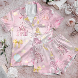 May include: Pink satin pyjama set with a princess and castle design. The set includes a short-sleeved button-up shirt and matching shorts. The design includes stars, crowns, and bows. The pyjamas are displayed on a pink fluffy surface.