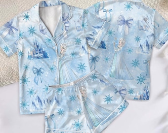 May include: Light blue pyjama set featuring a princess design, snowflakes, and bows. The set includes a short-sleeved button-up shirt and matching shorts. The pyjamas are decorated with a castle and mountain scene.