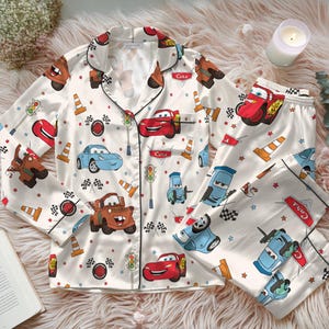 Può includere: Pigiama color crema a tema Cars. Il set comprende una camicia con bottoni e pantaloni, entrambi decorati con illustrazioni di auto colorate, coni stradali e bandiere a scacchi. La camicia ha finiture nere e un taschino sul petto. Il set pigiama è realizzato in materiale setoso.