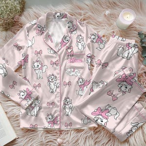 Puede incluir: Conjunto de pijama rosa claro con un estampado de gatos blancos de dibujos animados con lazos rosas. La parte superior de manga larga tiene un bolsillo en el pecho con el nombre "Anna" bordado. El conjunto es de material sedoso y está sobre una superficie rosa esponjosa.