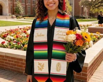 May include: A graduate in a black cap and gown smiles, holding a bouquet of flowers. She wears a colorful serape-style graduation stole with the name "Jaelene" and "Astravia Academy 2026" printed on it. The stole has a vibrant striped pattern.