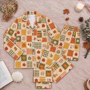 May include: A long-sleeved button-down pajama top and matching pants featuring a patchwork pattern of autumn-themed squares. The design includes illustrations of orange pumpkins, red and yellow maple leaves, acorns, and green plaid or polka dot motifs.