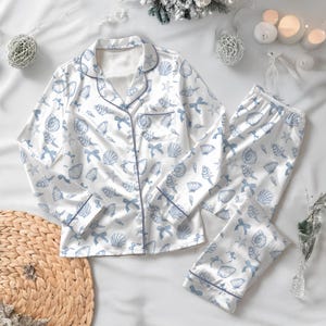 May include: White satin pajama set with a blue seashell and bow pattern. The long-sleeved top has a notched collar and a front pocket. The pants have an elastic waistband and a matching print. The set is trimmed with blue piping.