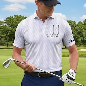 Op de afbeelding: Witte golfpolo met een geometrisch patroon, met golfclubafbeeldingen en de tekst "BEST DAD BY PAR". De shirt heeft een gepersonaliseerde naam op de mouw. De persoon houdt een golfclub vast op een groene golfbaan.