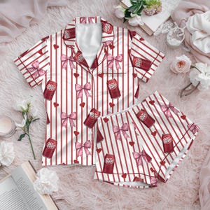 May include: A two-piece pajama set featuring a white and red striped pattern. The set includes a short-sleeved button-up shirt and shorts, adorned with pink bows, red hearts, and Dr. Pepper can graphics. The pajamas are made of a soft, silky material.