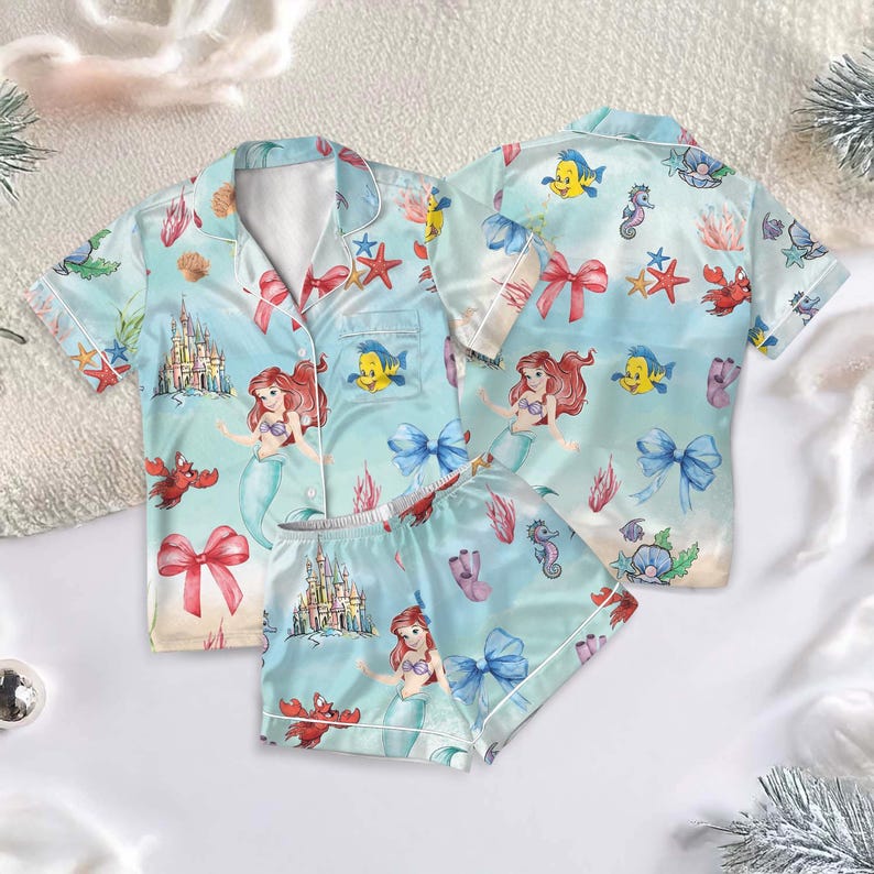 May include: Light blue pajama set featuring a mermaid theme. The set includes a button-down shirt and shorts, both adorned with illustrations of mermaids, castles, and sea creatures. The design incorporates bows, starfish, and other aquatic elements.