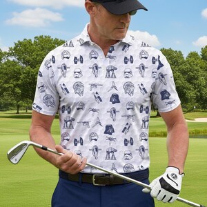 Op de afbeelding: Witte golfpolo met een marineblauw Star Wars-thema patroon. Het shirt heeft een herhalend ontwerp van verschillende Star Wars-personages en voertuigen. De persoon draagt een zwarte pet, een marineblauwe broek en een witte golfhandschoen.