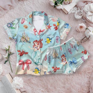 May include: Light blue pajama set featuring a print of Ariel from The Little Mermaid, castles, seahorses, and other sea creatures. The set includes a button-down shirt and matching shorts with white trim.