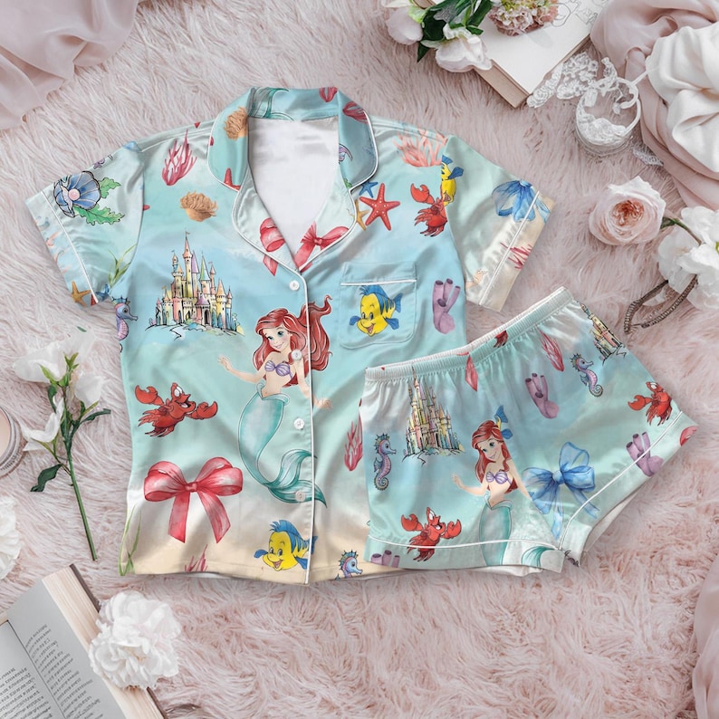 May include: Light blue pajama set featuring a print of Ariel from The Little Mermaid, castles, seahorses, and other sea creatures. The set includes a button-down shirt and matching shorts with white trim.