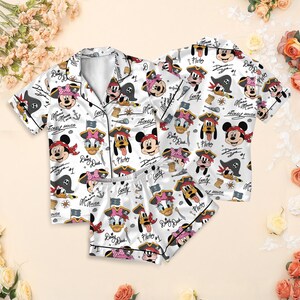 Könnte beinhalten: Weißes Pyjama-Set mit Piraten-Motiv von Disney-Figuren wie Mickey Mouse und Minnie Mouse. Das Set besteht aus einem kurzärmeligen Hemd mit Knöpfen und passenden Shorts. Der Druck zeigt Piratenhüte, Anker und Schatzkarten.