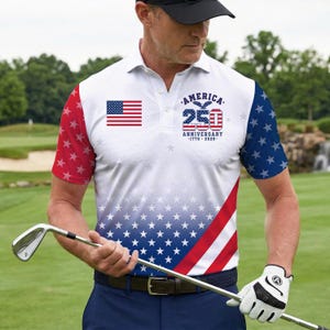Op de afbeelding: Witte poloshirt met een patriottisch ontwerp met de Amerikaanse vlag en sterren. Het shirt bevat de tekst "AMERICA 25TH ANNIVERSARY 1776-2026". De mouwen hebben rode en blauwe sterrenpatronen. De persoon draagt een zwarte pet en houdt een golfclub vast.