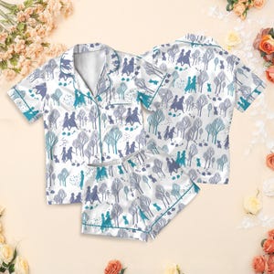 May include: White pajama set with a repeating pattern of figures, trees, and animals in shades of blue and purple. The set includes a short-sleeved button-up shirt and matching shorts, trimmed with teal piping. The fabric has a satin sheen.