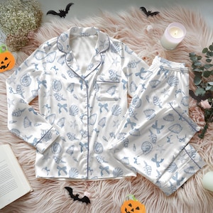 May include: White pajama set with a blue seashell and bow pattern. The long-sleeved top has a notched collar and a chest pocket. The pants have an elastic waistband. The set is trimmed with blue piping.