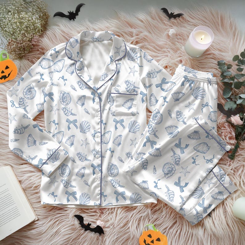 May include: White pajama set with a blue seashell and bow pattern. The long-sleeved top has a notched collar and a chest pocket. The pants have an elastic waistband. The set is trimmed with blue piping.
