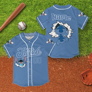 Peut inclure: Maillot de baseball bleu clair avec Stitch de Lilo & Stitch. Le maillot a des d&eacute;tails blancs, le nom "Stitch" et le num&eacute;ro "00" sur le devant. Le dos du maillot porte le nom "Name". Une batte et une balle de baseball sont en arri&egrave;re-plan.