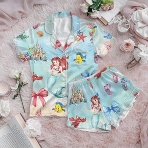 May include: Light blue satin pajama set featuring a mermaid theme. The set includes a button-down short-sleeved shirt and matching shorts. The design showcases Ariel, Flounder, a castle, and other sea creatures. The pajamas have white trim and a bow.