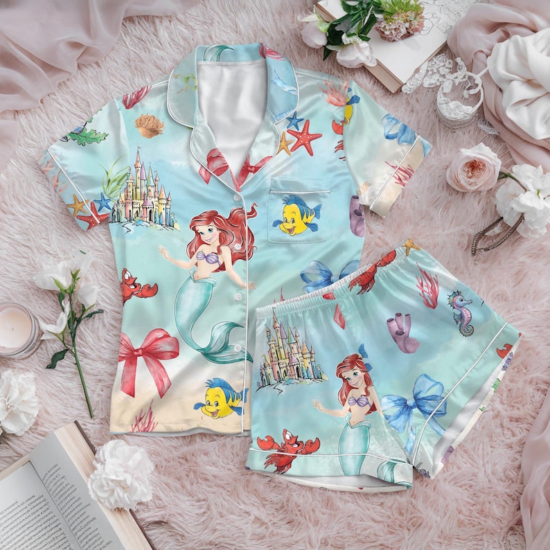 May include: Light blue satin pajama set featuring a mermaid theme. The set includes a button-down short-sleeved shirt and matching shorts. The design showcases Ariel, Flounder, a castle, and other sea creatures. The pajamas have white trim and a bow.
