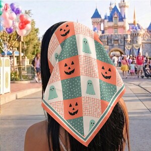 May include: A square head scarf featuring a patchwork pattern of orange jack-o'-lantern faces, light green ghosts, and white sections with small floral prints. The scarf has a grid-like orange border and is worn tied over dark hair.