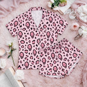 May include: A pink pajama set consisting of a short-sleeved button-down shirt with a chest pocket and matching shorts. The fabric features a repeating pattern of black leopard spots interspersed with black ghost-like face icons and white piping.