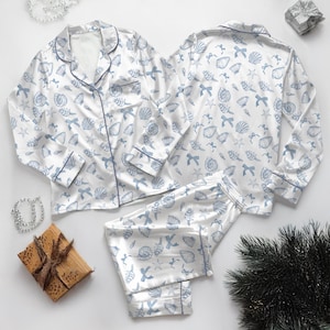 May include: White pajama set with a blue seashell and bow pattern. The set includes a button-down top with blue piping and long pants. The pajamas are made of a silky material.