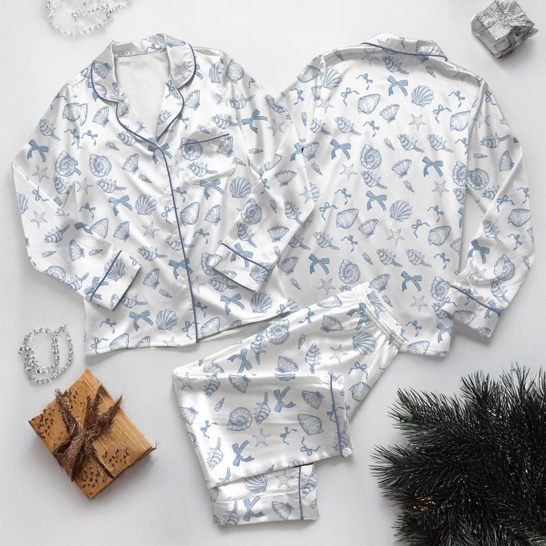 May include: White pajama set with a blue seashell and bow pattern. The set includes a button-down top with blue piping and long pants. The pajamas are made of a silky material.