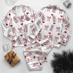 May include: White satin pajama set with a repeating pattern of Diet Coke cans, pink bows, and red hearts. The set includes a button-down top and pants, trimmed with red piping. A festive and fun sleepwear option.