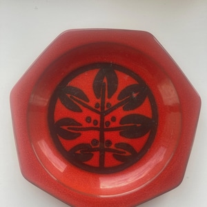 May include: A red, hexagonal dish with a dark brown, stylized floral design in the centre. The dish has a glossy finish and a slightly raised edge. The design features a central stem with leaf-like shapes.