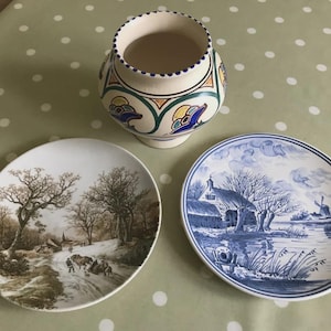 May include: A decorative ceramic vase with a cream base and a patterned design in blue, green, and yellow, accompanied by two plates. One plate depicts a winter scene in brown, the other a blue and white landscape.