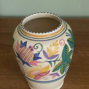 May include: A cream-coloured ceramic vase with a wide mouth and a rounded body. The vase is decorated with colourful floral patterns in shades of pink, blue, yellow, and green. The rim has a series of small, evenly spaced indentations.