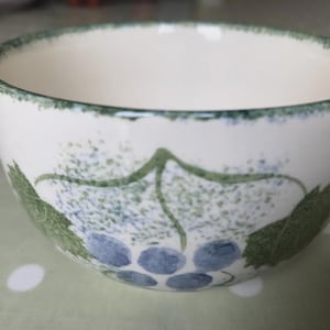 May include: A white ceramic bowl with a green rim and a grape design. The bowl features a hand-painted design of green leaves and blue grapes. The bowl is suitable for serving soups, cereals, or desserts.