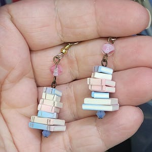 May include: Dangle earrings with miniature book stacks in pastel shades of pink, blue, and white. The earrings have gold-coloured hooks, pink beads, and blue crystal drops. A charming literary-themed accessory.