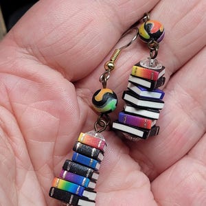 Puede incluir: Un par de pendientes únicos con pilas de libros en miniatura en colores del arcoíris. Cada pila de libros está acentuada con una cuenta transparente y una cuenta colorida. Los pendientes tienen alambres para las orejas de color dorado.