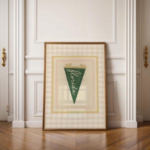 May include: A framed print featuring a green pennant with the word "Florida" in white script. The pennant is set against a plaid background with a gold border. The frame is a warm brown color. The print is displayed against a white wall.