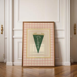 Op de afbeelding: Een ingelijste print met een groene wimpel met het woord "Custom" in witte letters. De wimpel staat tegen een crèmekleurige achtergrond met een gele rand, omlijst door een roze ruitpatroon. De lijst is warmbruin.