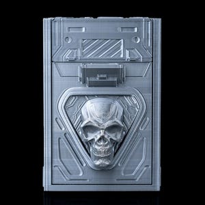 May include: A silver-colored box with a skull design. The box has a rectangular shape with a detailed, textured surface. The skull is centered on the front, within a triangular frame. The box has a latch closure.