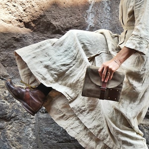 May include: A woman wearing a long, flowing white dress and brown boots holds a brown leather clutch with a buckle closure.