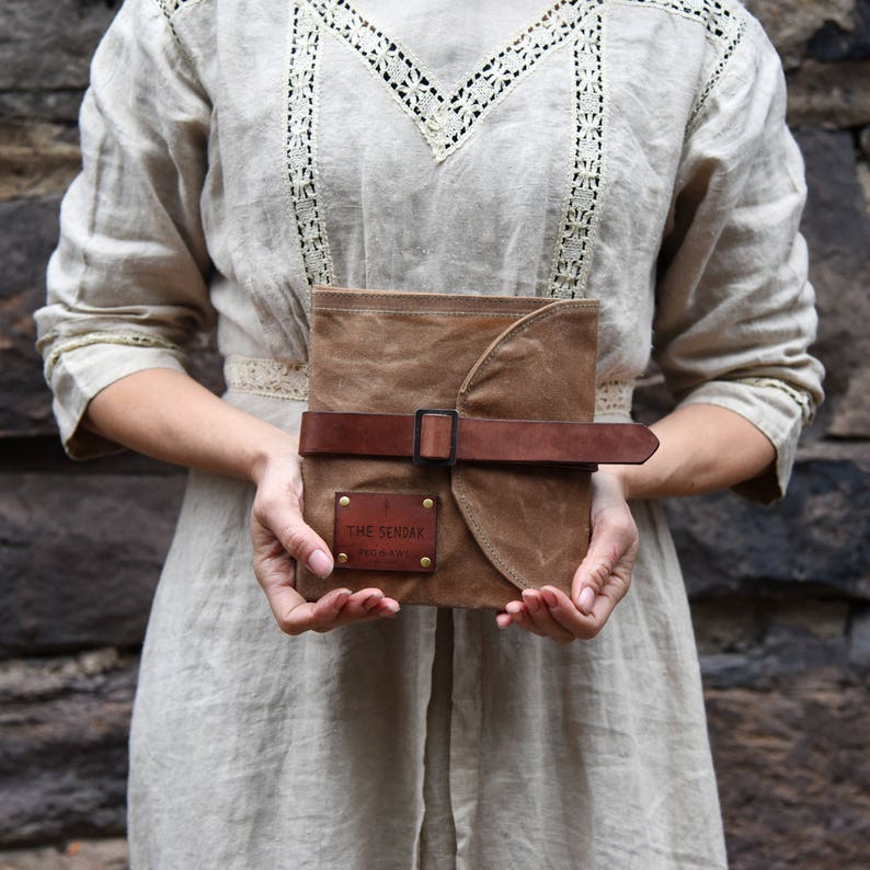 May include: A brown canvas journal with a leather strap and buckle closure. The journal has a brown leather patch with the text "THE SENDAK" embossed in gold.