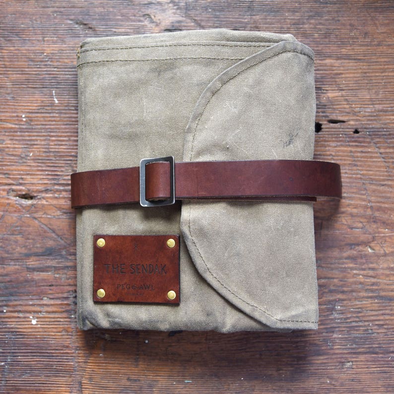 May include: A gray canvas roll-up pouch with a brown leather strap and buckle. The pouch has a brown leather label with the text "THE SENDAK PEG & AWL" in white.