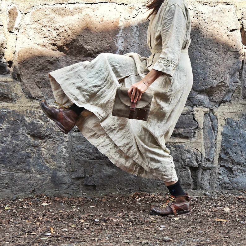 May include: A woman wearing a long, flowing white dress and brown boots holds a brown leather clutch with a buckle closure.