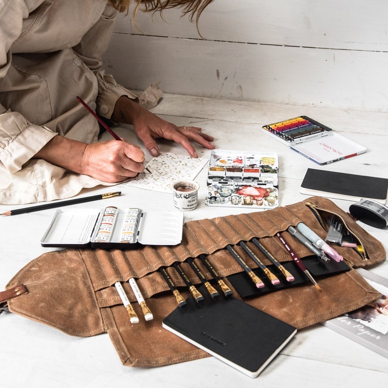 May include: A brown leather roll-up case with a variety of art supplies, including pencils, watercolor paints, and a small notebook. The case is open and the supplies are visible.