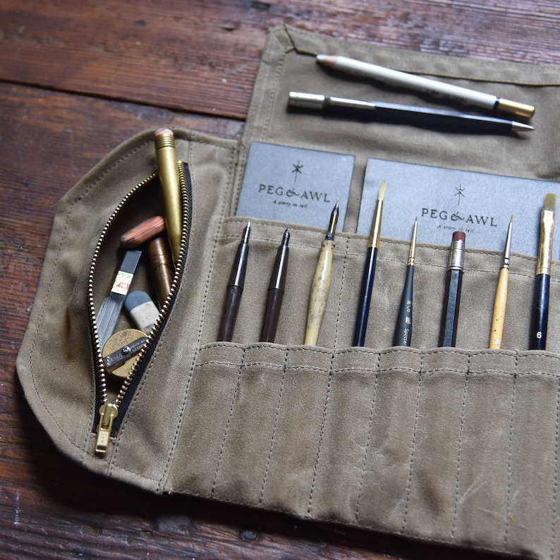 May include: A tan canvas roll-up case with a zipper closure. The case is filled with various art supplies, including pencils, brushes, and a pencil sharpener. The case has two labels that read "Peg & Awl, A Shop in NYC."