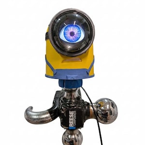 May include: A yellow and blue Minion-themed trailer hitch cover with a large eye. The eye is a lens with a blue iris and a black rim. The hitch is silver and black with the text "REESE TOWPOWER".