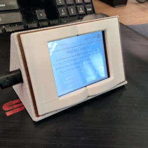 Puede incluir: Un dispositivo de pantalla digital blanco en forma de cuña con una pantalla rectangular que muestra el texto Psalms 140:1. El dispositivo cuenta con una capa interior marrón, un cable de alimentación negro conectado al lado y un marco blanco.